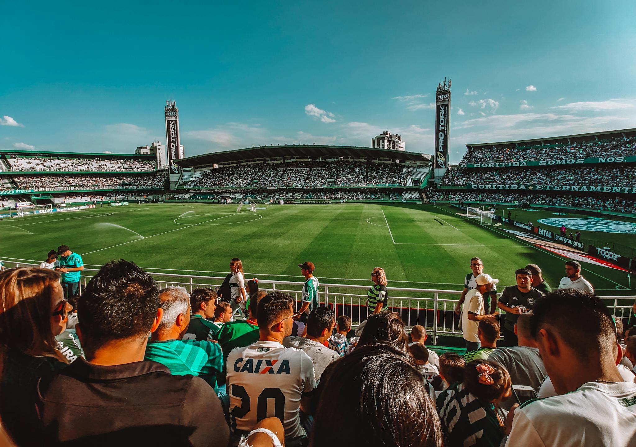 Fans enjoy a vibrant soccer match in sunny outdoor stadium with a large crowd and blue sky.